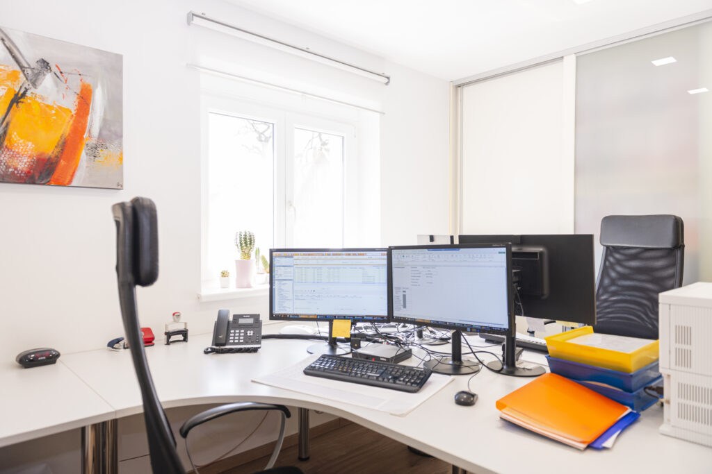 Arbeitsumfeld Steuerberatungskanzlei - Helles, modernes Büro der Steuerberatungskanzlei mit mehreren Monitoren, Schreibtisch, Bürostuhl und farbigen Aktenordnern. Am Fenster stehen kleine Pflanzen, ein Bild in Orange- und Grautönen schmückt die Wand.