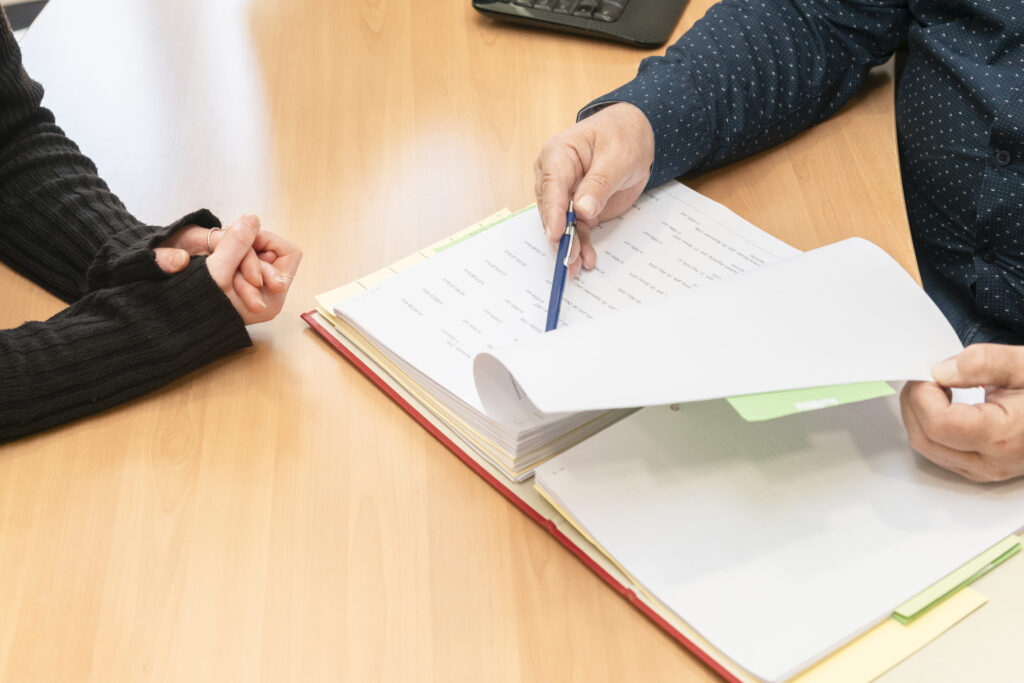 Genaue Arbeit in der Steuerberatungskanzlei - Zwei Personen bei einer Besprechung an einem Holztisch: Eine Person blättert mit einem Stift in einem Aktenordner, die andere hört aufmerksam zu.